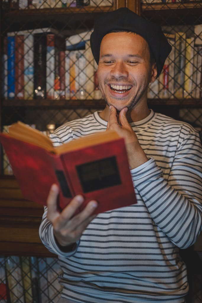 A cheerful man in a striped shirt reads a book in a cozy library setting.